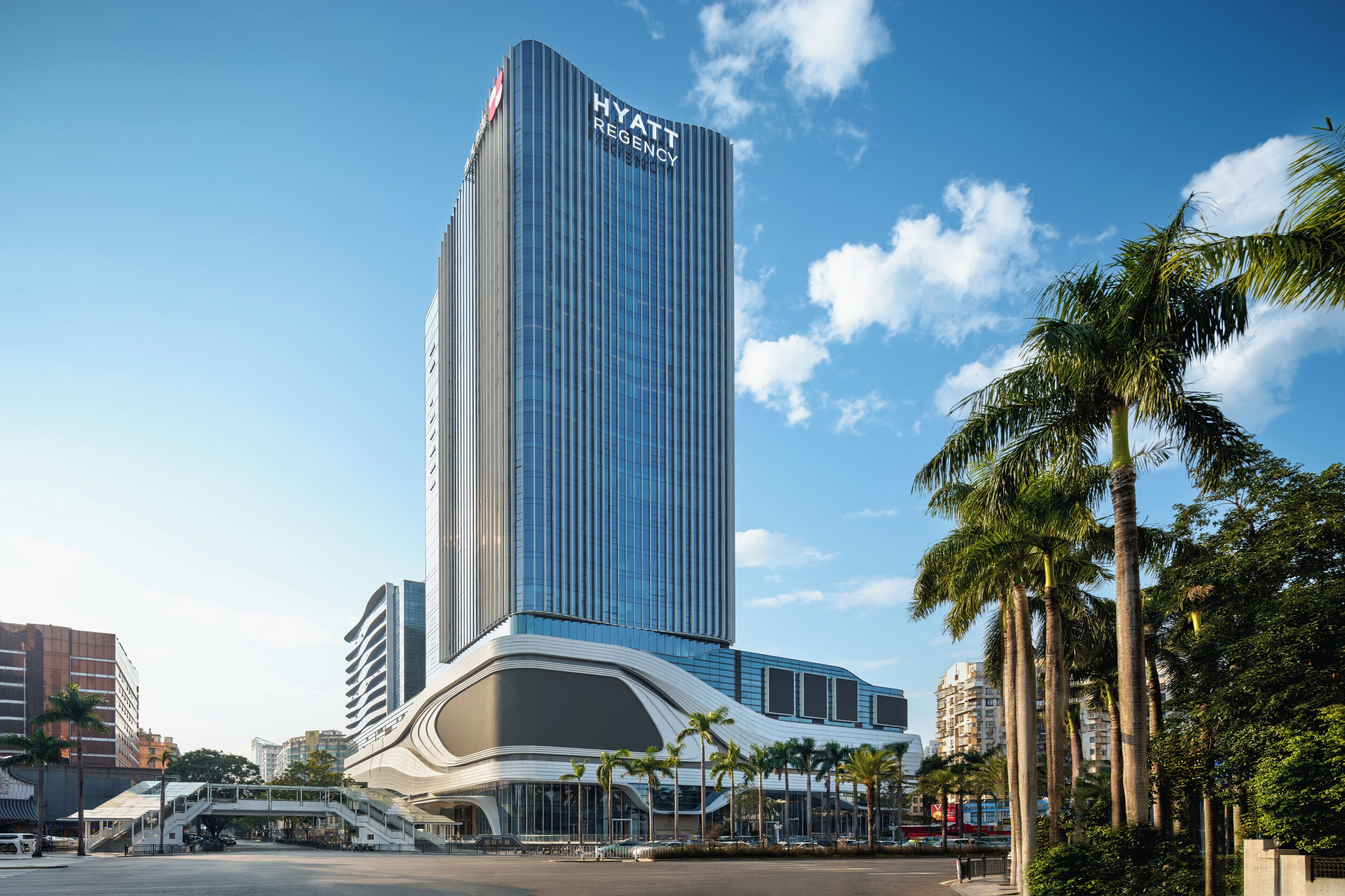 A modern high-rise hotel with a prominent 'Hyatt Regency' sign, a curved base structure, and palm trees in the foreground.
