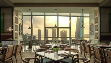 An indoor dining area with tables and chairs, large windows with a city view, and pendant lighting.