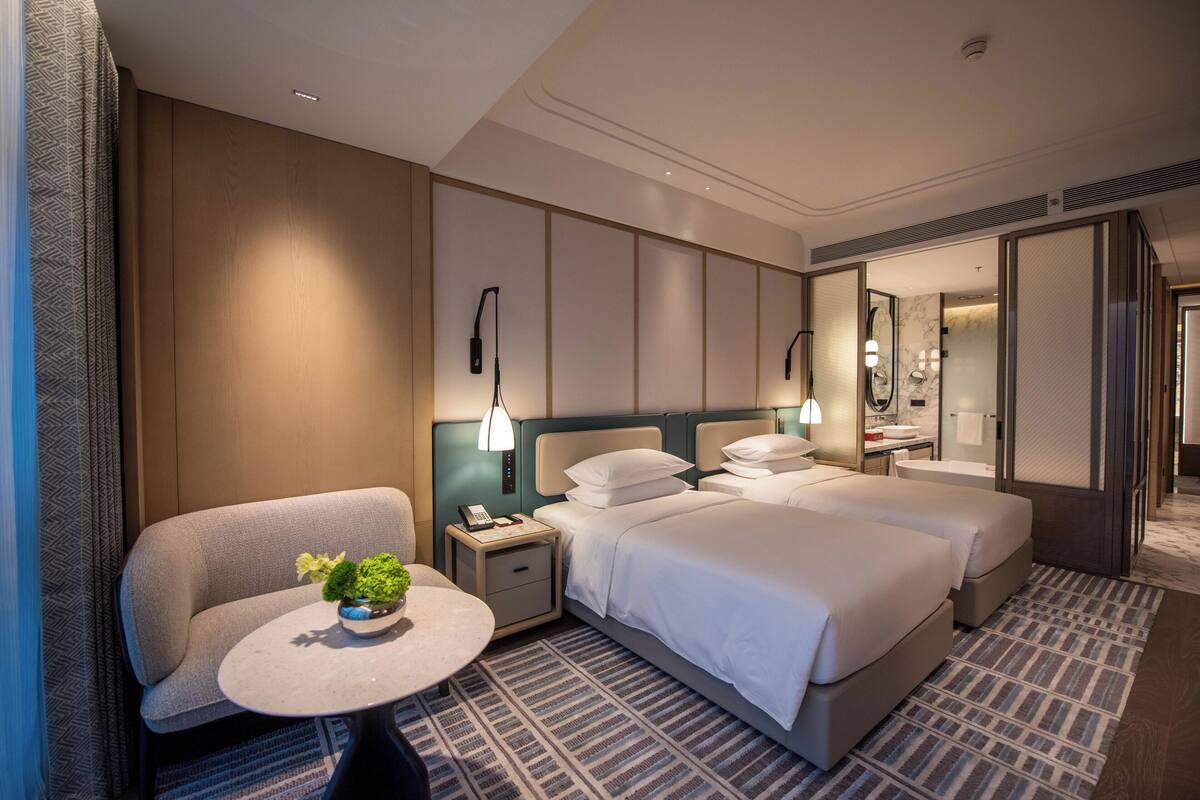 Una habitación de hotel con cama, mesitas de noche, una silla, una pequeña mesa con una planta y un espejo.