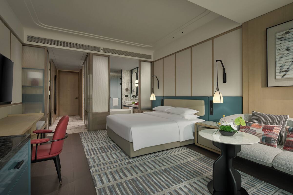 Habitación de hotel con cama, escritorio, silla, sofá, una mesita con planta y televisor montado en la pared.