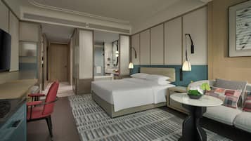 Habitación de hotel con cama, escritorio, silla, sofá, una mesita con planta y televisor montado en la pared.