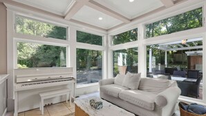 Ein geräumiges Wohnzimmer mit einem weißen Klavier, einem beigefarbenen Sofa und großen Fenstern mit Blick ins Freie.