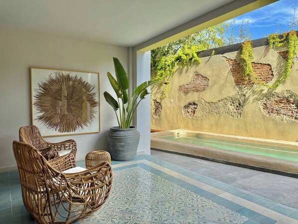 A room with a large window overlooking a pool, wicker furniture, a potted plant, and a wall with a textured mural. - A pasos del Centro Historico, Bares, con Alberca (Mazatlán)