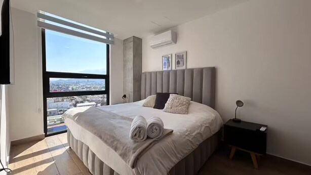 A modern bedroom with a large bed, a view of the city through floor-to-ceiling windows, and a small bedside table with a lamp. - Chic 3BR Prime Spot - 12 guests (Monterrey)