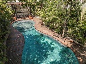 A curved swimming pool with wooden decking, surrounded by lush greenery and a small outdoor seating area with chairs and tables. - Exclusive Condo - Free Beach Pass & Bikes & Private Pool (Tulum)
