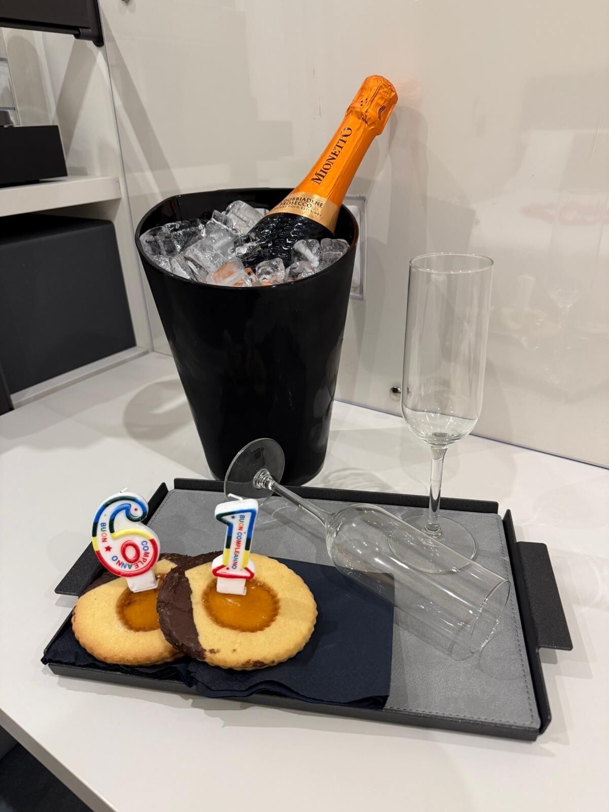 Decoración para una celebración de cumpleaños con una botella de champán Mionetto en un cubo negro, una copa de champán y dos galletas con velas.
