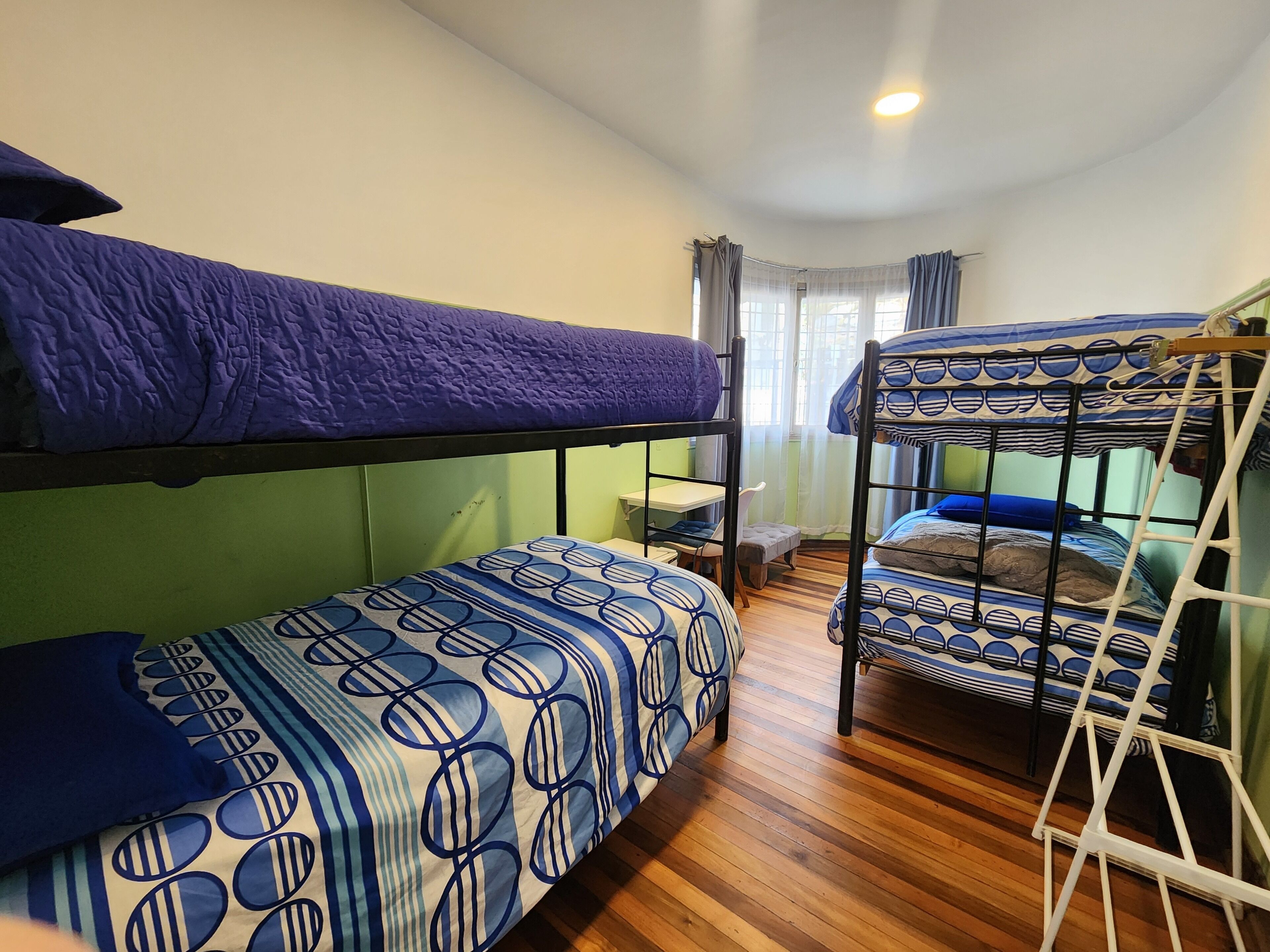 A dormitory room with bunk beds, a small desk, and a window with curtains.