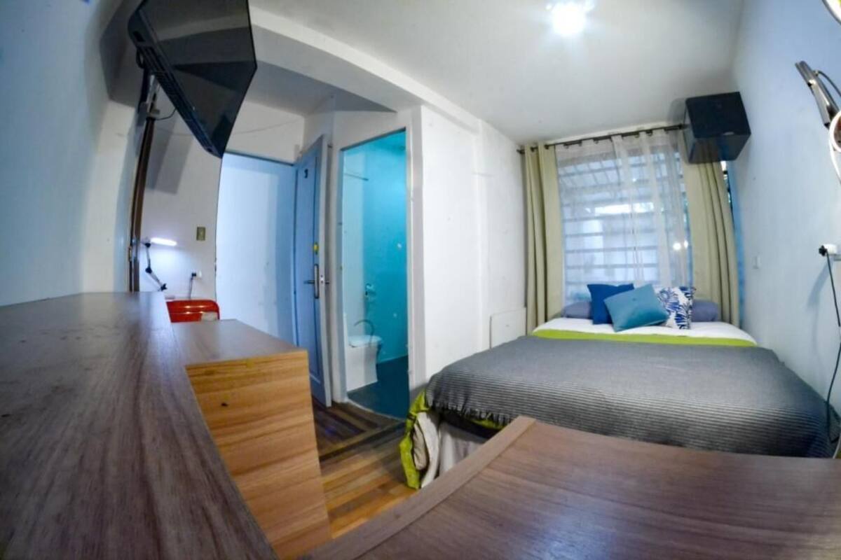 Une chambre avec un bureau en bois, un lit recouvert d’une couverture verte et blanche, une salle de bain avec une porte bleue et un plafond blanc.