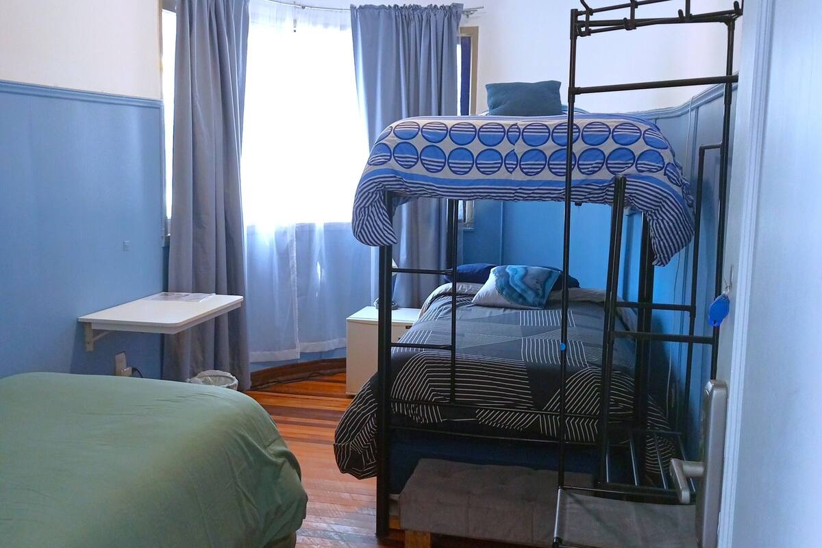 Une chambre avec un lit superposé, un lit simple, une petite table et une fenêtre avec des rideaux.