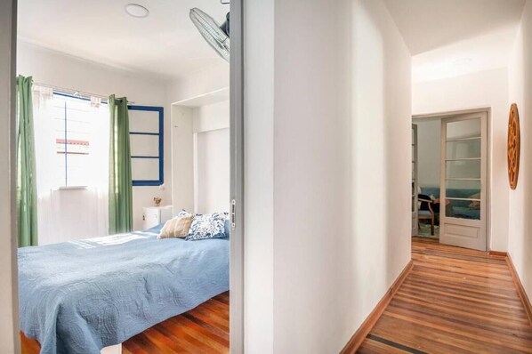 A bedroom with a bed, a window with curtains, a fan, and a view of a balcony.