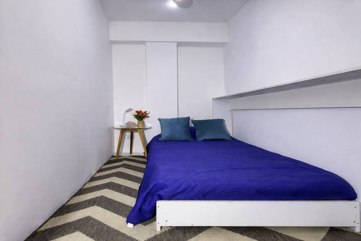 Une petite chambre à coucher moderne, avec un lit blanc, une literie bleue et une petite table sur laquelle se trouvent une lampe et des fleurs.