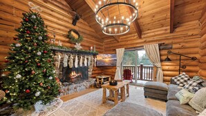Ein gemütliches Blockhaus-Interieur mit geschmücktem Weihnachtsbaum, Kamin, Sofa und Blick ins Freie.