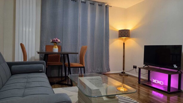 Una sala de estar moderna con un sofá, una mesa de centro de vidrio, un televisor en un mueble, una lámpara y un área de comedor con sillas y una mesa.