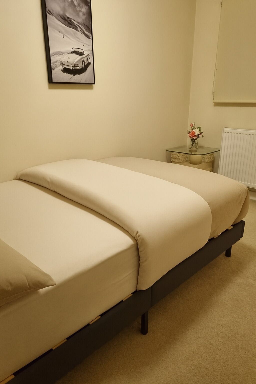 A bed with a dark frame and beige bedding in a room with a plain wall and a small table with a vase of flowers.