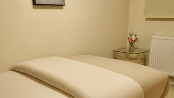 A bed with a dark frame and beige bedding in a room with a plain wall and a small table with a vase of flowers.
