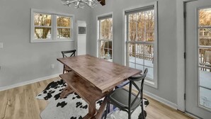 A dining area with a wooden table, chairs, a chandelier, and large windows with a view of a snowy landscape. - Walk to Slopes-Mountain Views-Firepit-Sleeps 10 (Beech Mountain)