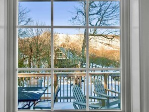 Vista a través de una ventana que muestra una terraza de madera, mobiliario exterior y una casa al fondo.