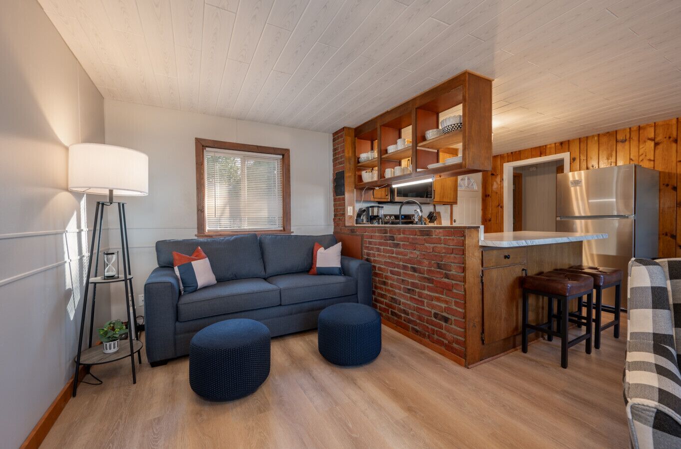 Un espacio habitable compacto con sofá, otomanas, una pequeña zona de cocina con refrigerador y una ventana con persianas.