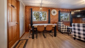 Un acogedor interior de cabaña con una mesa de comedor de madera, cuatro sillas, un sofá a cuadros y un televisor grande.