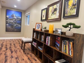 A room with a wooden floor, a bookshelf with books, framed pictures on the wall, a chair, and a window.