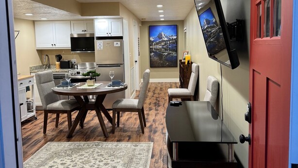 A modern kitchen with white cabinets, a dining area with a round table and chairs, a flat-screen TV mounted on the wall, and a red door. - Stay cool in Basement apartment close to Mountain Trails (Sandy)