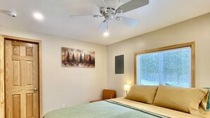 A bedroom with a bed, a ceiling fan, a window with blinds, a door, and a painting on the wall. - Moonlight Cottage | McKinney Lake Cottages | One Bedroom | One Bath (Grand Rapids)