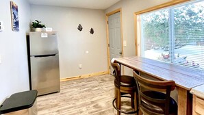 A compact kitchen with a refrigerator, a wooden dining table with two chairs, and a window with blinds. - Moonlight Cottage | McKinney Lake Cottages | One Bedroom | One Bath (Grand Rapids)