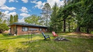A log cabin with a grassy yard, a red chair, a blue chair, and a fire pit. - Moonlight Cottage | McKinney Lake Cottages | One Bedroom | One Bath (Grand Rapids)