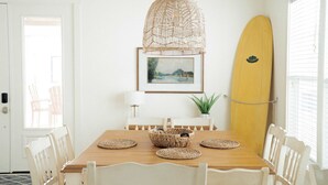 Un comedor con una mesa de madera, sillas blancas, una lámpara colgante tejida, un cuadro enmarcado y una tabla de surf apoyada contra la pared.
