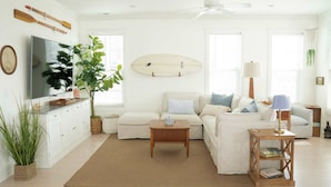 Una sala de estar con un sofá blanco, una mesa de centro, una tabla de surf en la pared y un televisor sobre un mueble.
