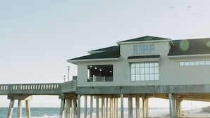 Un edificio costero con balcón, un muelle que se adentra en el mar y un cielo despejado.