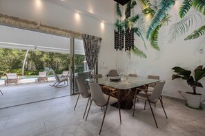 A dining area with a glass table surrounded by chairs, a large potted plant, and a view of the outdoors.