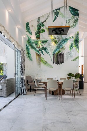 A dining area with a modern table and chairs, a hanging light fixture, and a wall mural of tropical leaves.