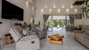 Un salon moderne doté d’un téléviseur à écran plat, de canapés inclinables, d’une table basse en bois et offrant une vue sur l’extérieur grâce à de grandes fenêtres.