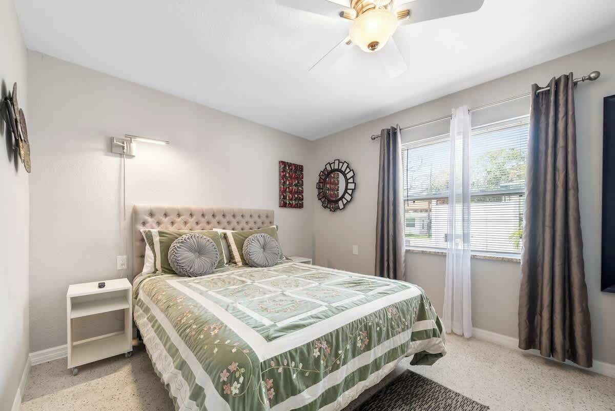 Quarto com cabeceira estofada, cama com colcha florida, luminária de chão, espelho decorativo e janela com cortinas.