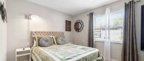 Quarto com cabeceira estofada, cama com colcha florida, luminária de chão, espelho decorativo e janela com cortinas.