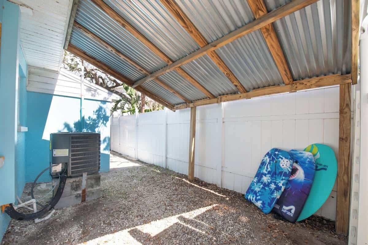 A covered outdoor area with a gravel floor, a white wall, and a blue wall. There is a black air conditioning unit and a surfboard leaning against the wall.
