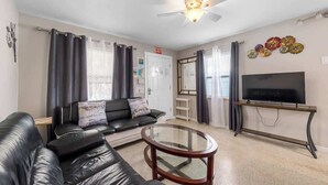 A living room with a black leather sofa, a glass coffee table, a ceiling fan, a TV on a wooden stand, and a door with an 'EXIT' sign. - 3 Min Walk to Beach | Modern Coastal Retreat (Indian Rocks Beach)