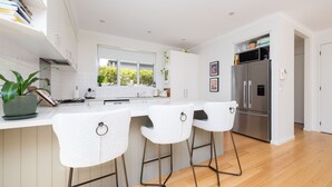 A modern kitchen with white cabinetry, a central island with bar stools, a stainless steel refrigerator, and a window with a view of greenery. - Pet Friendly - Evergreen by the Bay (Sandringham)