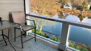 Uma varanda com mesa e cadeira, com vista para um lago e casas e árvores ao fundo.