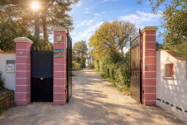 Une entrée fermée par un portail, sur laquelle est apposée une plaque indiquant « LA BASTIDE », et un sentier qui mène à une zone boisée.