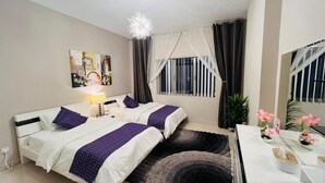 Habitación de hotel con dos camas, un escritorio, una silla, una lámpara, una ventana con cortinas, una planta y un cuadro en la pared.