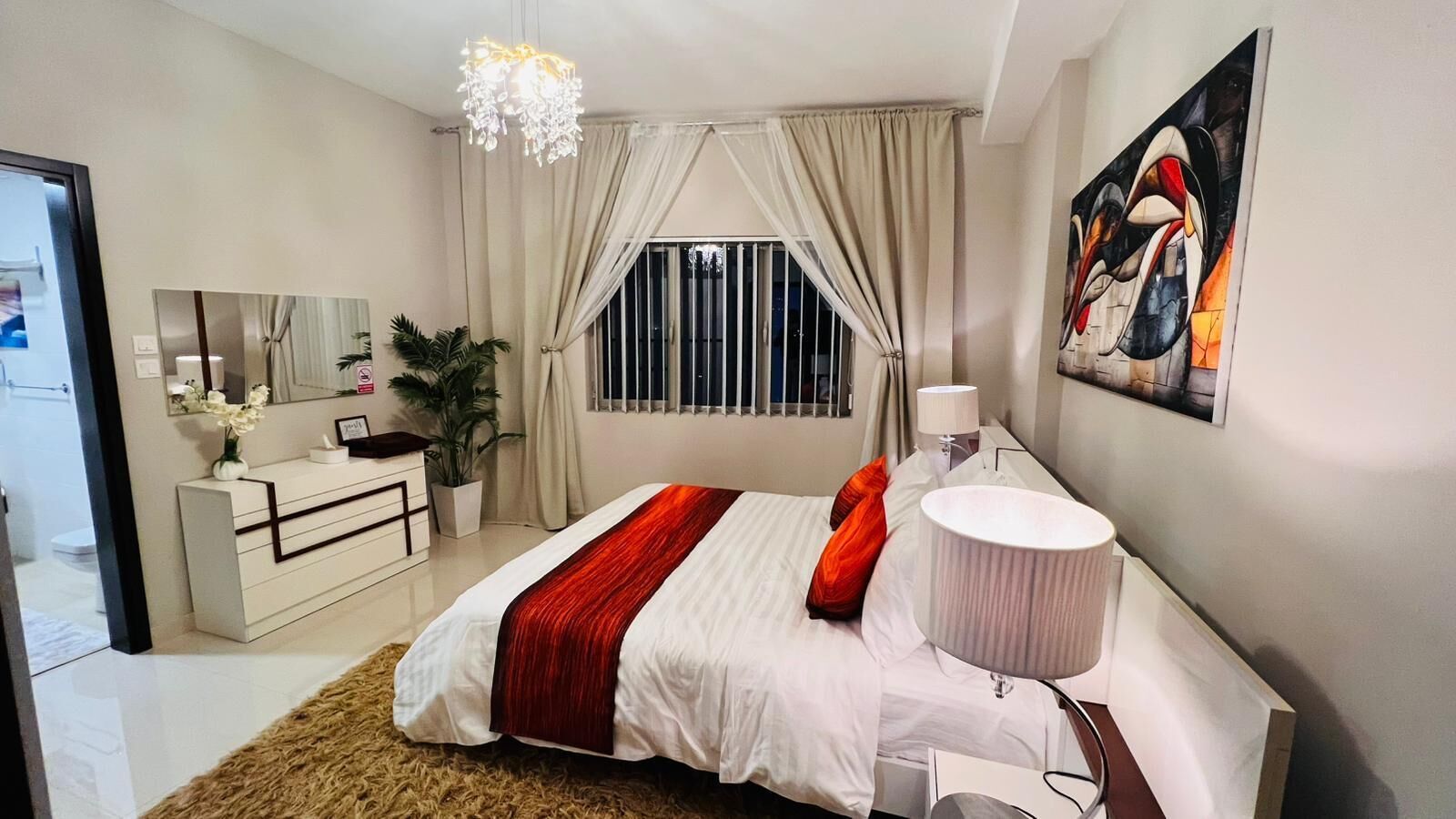 A modern bedroom with a large bed, a bedside table, a lamp, a mirror, a window with curtains, and a painting on the wall.