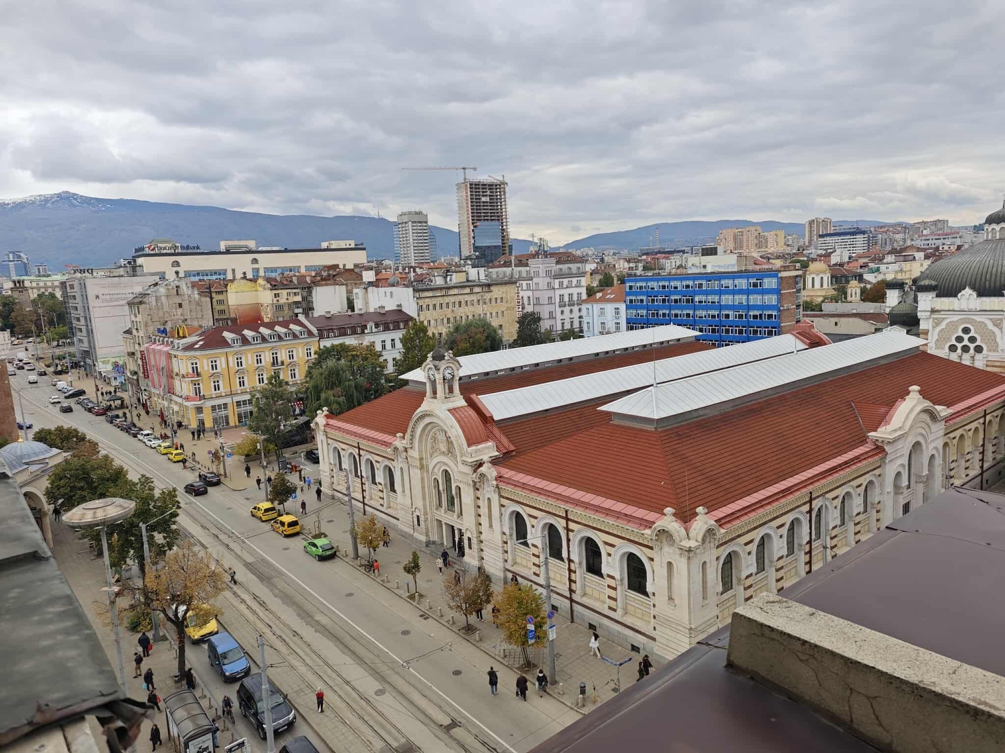 A historic building with a red roof and classical architecture, surrounded by modern buildings and a busy street.
