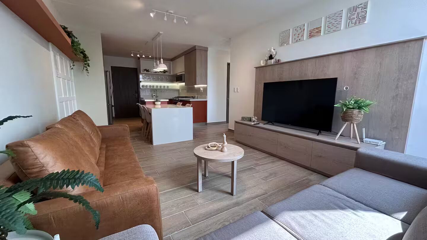 Ett modernt vardagsrum med en brun lädersoffa, en grå soffa, ett litet runt soffbord och en platt-TV som är monterad på en väggpanel av trä.