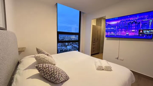 Ein Hotelzimmer mit einem großen Bett, einem an der Wand befestigten Fernseher und Blick auf eine Stadtlandschaft durch ein Fenster.