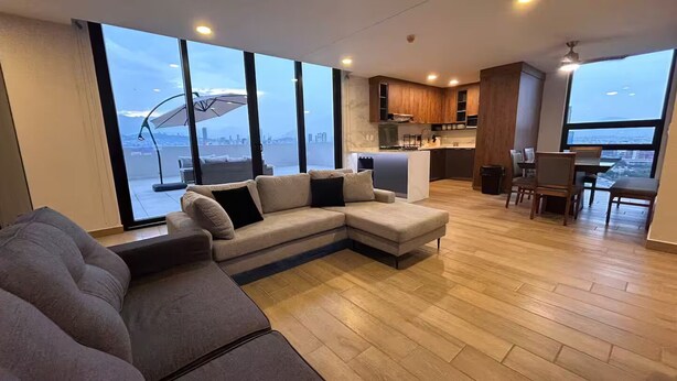 Ein modernes Wohnzimmer mit einem großen Ecksofa, einem Essbereich mit Tisch und Stühlen, einer Küche mit Holdekor und großen Fenstern, die einen Blick auf die Stadt ermöglichen.