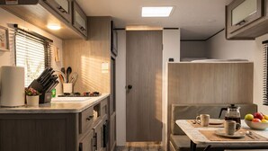 A compact RV kitchen with wooden cabinets, a countertop with utensils, and a dining area with a table and chairs.