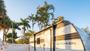 A Dutschmen RV parked in a backyard with artificial grass, turquoise chairs, and a small table with a book and a lantern.