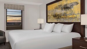 A hotel room with a large bed, a nightstand with a lamp, a chair, and a large window with a view of the sunset. - Family-Friendly 2BR Suite at Ocean Walk - Beach & Pools Await (Daytona Beach)